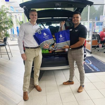 Two men holding boxes standing by an open car trunk in a showroom.